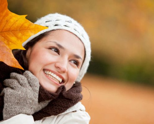 Kobieta uśmiecha się, trzymając jesienny liść i zachwyca odmłodzoną twarzą po zabiegach kosmetycznych wykonanych jesienią.