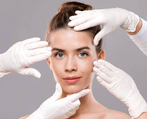 Twarz kobiety, której dotykają cztery dłonie w jednorazowych, białych rękawiczkach – kosmetolożek z salonu kosmetycznego, które wyjaśniają swojej klientce, jak można poprawić owal twarzy.
