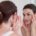 Ciemnowłosa kobieta w białej koszulce na ramiączkach przegląda się w lustrze i patrzy na swoją gładką twarz zadowolona, że zdecydowała się na zabieg radiofrekwencja mikroigłowa na oczy i twarz.